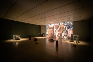 In einer schwach beleuchteten Galerie stehen verstreut moderne Skulpturen auf dem Boden und ein großer Bildschirm zeigt ein abstraktes Bild mit Industriemotiven.