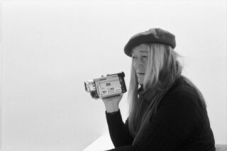 Eine ältere Frau mit Baskenmütze und Schal hält eine Videokamera in der Hand und blickt zur Seite auf einen schlichten Hintergrund.