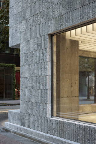 Nahaufnahme einer modernen Gebäudeecke mit strukturierten Betonwänden und einem großen Fenster, das den Blick auf ein minimalistisches Interieur freigibt.