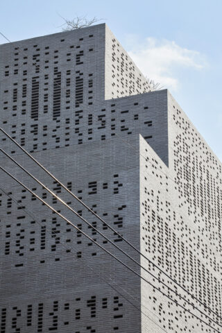 Ein abgestuftes modernes Gebäude mit kleinen rechteckigen Fenstern in einem Gittermuster, von unten gesehen vor einem blauen Himmel mit sichtbaren Stromleitungen im Vordergrund.
