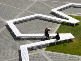 Zwei Personen sitzen auf einer weißen, kantigen, modularen Bank auf einem geometrischen Platz mit Gras und gemusterten Pflastersteinen.