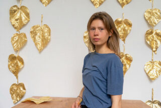 Eine Frau in einem blauen Hemd sitzt an einem Holztisch vor einer weißen Wand, die mit hängenden goldfarbenen Blättern verziert ist.