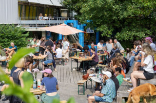 Die Menschen sitzen an Tischen im Freien unter einem großen Sonnenschirm, essen und unterhalten sich in einem belebten Innenhof, der von Grünflächen und Gebäuden umgeben ist.