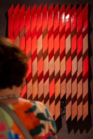 Eine Person steht vor einem geometrischen Kunstwerk aus diagonalen roten und braunen Paneelen, die in vertikalen Reihen an einer dunklen Wand angeordnet sind.