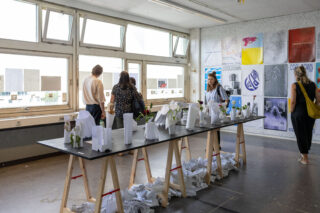 Die Besucher betrachten Kunst- und Designprojekte, die auf einem Tisch in einem hell beleuchteten Raum ausgestellt sind, an dessen Wänden und Fenstern Plakate und Papiermodelle hängen.