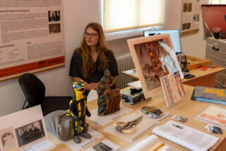 Eine Frau sitzt an einem Schreibtisch, auf dem verschiedene Kunstgegenstände, Fotos und Dokumente ausgestellt sind, in einem gut beleuchteten Raum, an dessen Wänden Informationsplakate hängen.