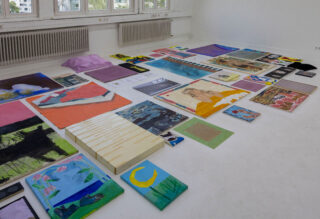 In einem hell beleuchteten Raum mit großen Fenstern und Heizkörpern an der Wand sind verschiedene farbenfrohe Gemälde und Kunstwerke auf einem weißen Boden ausgestellt.