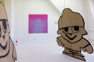 Eine Kunstgalerie mit zwei großen Comic-Ausschnitten im Vordergrund und einem Gemälde mit violettem Farbverlauf an der Rückwand. Der Raum ist hell und minimalistisch gestaltet.