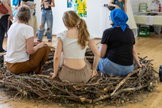 Drei Frauen sitzen in einem großen Nest aus Ästen auf einem Holzboden in einem Innenraum, im Hintergrund sind Kunstwerke und andere Menschen zu sehen.