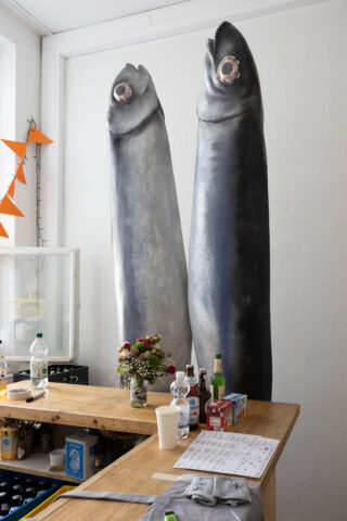 In einem hell erleuchteten Raum stehen zwei große Fischskulpturen hinter einer Holztheke, auf der Getränke, Blumen, Pappbecher und Zeitungen liegen.
