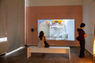 Zwei Personen betrachten eine Videoprojektion in einer Kunstgalerie; eine sitzt auf einer Bank, die andere steht rechts davon und blickt auf die Leinwand, auf der eine Bettszene mit deutschen Untertiteln zu sehen ist.