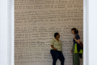 Zwei Personen stehen vor einer großen Wand, die mit handgeschriebenem deutschen Text in schwarzem Filzstift bedeckt ist, der fast den gesamten sichtbaren Raum ausfüllt, und unterhalten sich.
