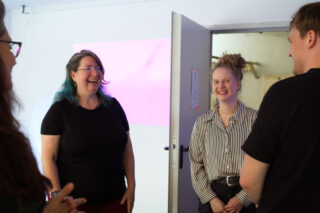 Vier Personen stehen in einem Raum nahe einer offenen Tür, unterhalten sich und lächeln. An der Wand hinter ihnen ist ein violettes Licht zu sehen.