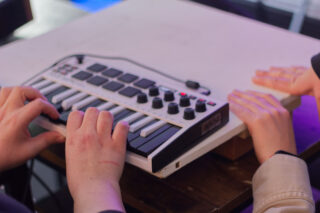 Zwei Personen benutzen einen kleinen MIDI-Keyboard-Controller auf einer weißen Plattform, wobei eine Person die Tasten drückt und die andere die Hände in der Nähe ruhen lässt.