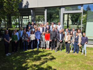 Eine Gruppe von etwa 35 Personen steht draußen auf dem Rasen vor einem modernen Gebäude mit großen Fenstern und posiert im Sonnenlicht für ein Gruppenfoto.