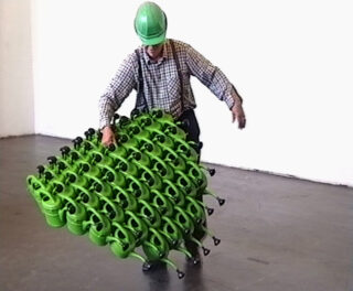 Eine Person mit grünem Schutzhelm und kariertem Hemd hält ein großes, gitterartiges Gebilde aus zahlreichen miteinander verbundenen grünen Gießkannen.