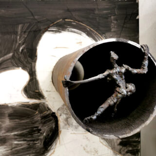 An der Öffnung eines verrosteten Rohrs ist eine Metallskulptur in Form einer menschlichen Figur befestigt, im Hintergrund sind abstrakte Schwarz-Weiß-Kunstwerke zu sehen.