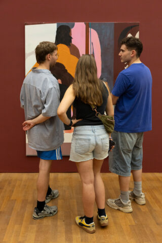 Drei Personen stehen in einer Kunstgalerie vor einem farbenfrohen abstrakten Gemälde und unterhalten sich.