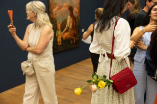 Eine Gruppe von Menschen steht in einer Kunstgalerie; eine Frau hält einen Fächer in der Hand, eine andere hat eine rote Handtasche und Blumen, die an ihrer Hand hängen. Im Hintergrund hängt ein Gemälde an der Wand.
