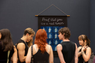 Fünf Personen stehen vor einem Kunstwerk mit Fotos von Händen und der Aufschrift „Der Beweis, dass du ein echter Mensch bist“ an einer dunklen Wand in einer Galerie.