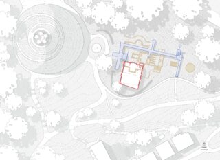 Lageplan, der ein Gebäude mit blau und rot umrandeten Bereichen zeigt, umgeben von Bäumen, Wegen, einer kreisförmigen Anlage und mehreren rechteckigen Bauwerken im unteren Bereich.