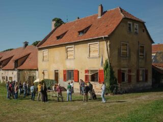 An einem sonnigen Tag versammelt sich eine Gruppe von Menschen vor einem großen gelben Haus mit vernagelten Fenstern und roten Fensterläden.