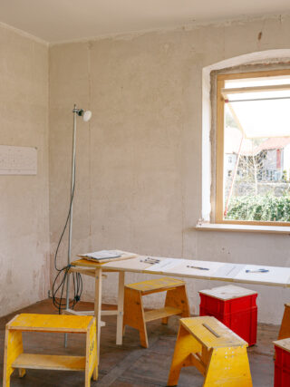 Ein schlichter Raum mit unverputzten Wänden, einem Holztisch, gelben Hockern, einer roten Kiste, Papieren, Stiften und einer Stehlampe neben einem Fenster, durch das Tageslicht hereinfällt.