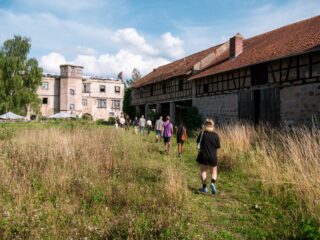 Eine Gruppe von Menschen geht unter einem teilweise bewölkten Himmel durch hohes Gras auf alte, verwitterte Gebäude zu.