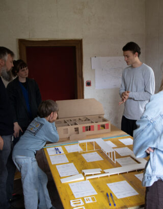 Fünf Personen sitzen um einen Tisch herum, auf dem Architekturmodelle, Papiere und Stifte liegen; eine Person hält eine Präsentation, während die anderen in einem Raum mit schlichten Wänden und einer roten Türöffnung zuschauen und zuhören.
