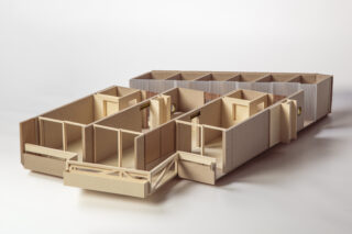 Ein Architekturmodell aus Pappe zeigt den Grundriss eines einstöckigen Gebäudes mit mehreren miteinander verbundenen Räumen und Fluren vor einem schlichten weißen Hintergrund.