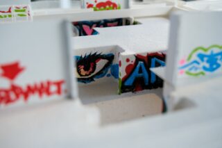 Nahaufnahme eines weißen Architekturmodells mit bunten Graffiti, darunter ein detailreiches Auge und kräftige blaue Buchstaben, die auf die Innenwände gemalt sind.