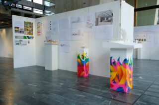 Architekturmodelle und Entwurfszeichnungen werden in einem modernen Ausstellungsraum mit grauem Fliesenboden auf weißen Wänden und bunten Sockeln präsentiert.