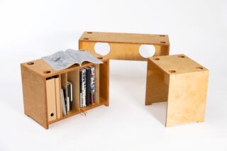 Es werden drei modulare Holzmöbelstücke gezeigt: Eines dient zur Aufbewahrung von Büchern und Papieren, eines fungiert als Bank und eines ist ein kleiner Beistelltisch. Alle weisen auf ihren Oberflächen kreisförmige Aussparungen auf.