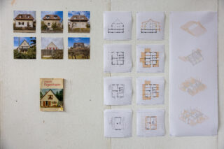 An einer weißen Wand sind Fotos von Häusern, ein Buch mit dem Titel „Mein Eigenheim“ sowie verschiedene architektonische Grundrisse und Zeichnungen ausgestellt.