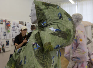 In einem Studio präsentieren Schaufensterpuppen Daunenjacken mit Naturmotiven; im Hintergrund unterhalten sich zwei Personen vor einer Wand mit Referenzbildern.