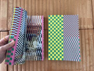 Eine Hand blättert in einem bunten Buch mit geometrischen Mustern auf einer strukturierten Ziegeloberfläche; ein weiteres Buch mit einem ähnlichen Einband liegt geschlossen daneben.