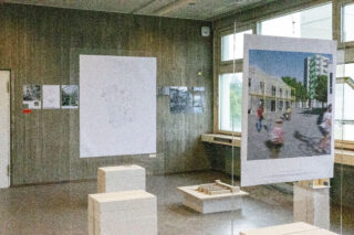 In einem Raum sind Architekturzeichnungen, Modelle und eine großformatige Darstellung eines Gebäudes auf transparenten Platten ausgestellt, während Tageslicht durch die Fenster hereinfällt.