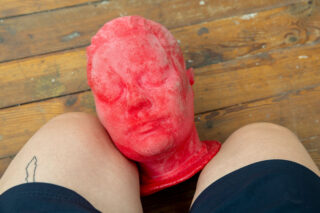 Ein roter, modellierter Kopf ruht auf den nackten Oberschenkeln einer Person, die schwarze Shorts trägt und auf einem Holzboden sitzt.