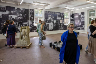 Die Besucher betrachten Kunstinstallationen und Schwarz-Weiß-Fotocollagen an den Wänden eines hell beleuchteten Galerieraums mit großen Fenstern und Fliesenboden.