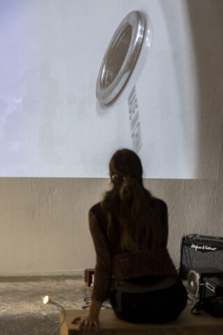 Eine Person sitzt auf einer Bank und blickt auf ein großes, an die Wand projiziertes Bild einer Waschmaschine; auf dem Boden in der Nähe sind Audiogeräte zu sehen.