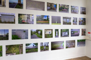 An einer weißen Wand ist ein Raster aus Farbfotos zu sehen, die verschiedene Außenaufnahmen zeigen, darunter Straßen, Gebäude, Zäune, Gras und Mauern; auf jedem Foto ist ein rechteckiger weißer oder grauer Fleck zu erkennen.