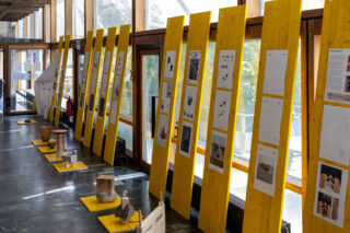 In einem sonnendurchfluteten Flur sind auf gelben Holztafeln Skizzen, Fotos und Texttafeln ausgestellt; vor jeder Tafel stehen verschiedene kleine Skulpturen auf dem Boden.