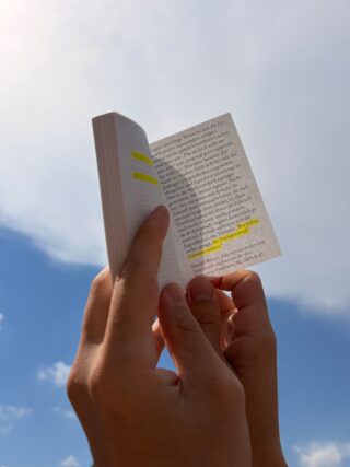 Eine Person hält ein kleines Buch mit gelb markiertem Text vor einen hellen Himmel mit vereinzelten Wolken.