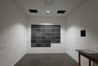 Ein kleiner Raum mit Fliesenboden und weißen Ziegelwänden, an dessen Rückwand ein Raster aus schwarzen Quadraten zu sehen ist und an dessen rechter Wand ein Tisch mit einem kleineren, gerahmten schwarzen Quadrat steht.