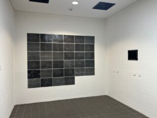 Ein weiß gefliester Raum mit einem Raster aus schwarzen Fliesen an einer Wand, einem kleinen schwarzen rechteckigen Display an einer anderen Wand und einem Fliesenboden.
