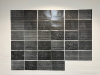 Ein Raster aus 33 rechteckigen schwarzen Paneelen, die an einer weiß gefliesten Wand angeordnet sind; in der rechten unteren Ecke fehlen drei Paneele.