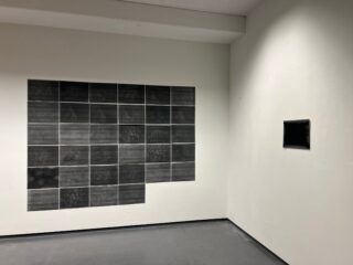 An einer weißen Wand ist ein Raster aus dunklen rechteckigen Paneelen angeordnet; an einer angrenzenden Wand in einem weitgehend leeren Raum ist ein einzelnes, kleineres dunkles Paneel zu sehen.