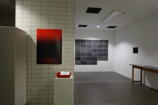 Ein mit Fliesen ausgekleideter Raum mit einem roten Schild an der linken Wand, einem kleinen weißen Sockel mit einem Tablett, einer Kunstinstallation aus schwarzen Fliesen an der gegenüberliegenden Wand und einem langen Tisch auf der rechten Seite.
