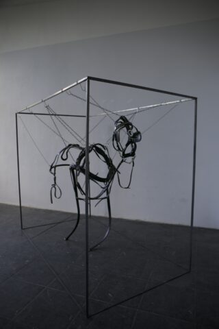 Eine minimalistische Pferdeskulptur aus schwarzen Riemen und Metallketten ist in einem karg gestalteten Galerieraum in einem kubischen Metallrahmen aufgehängt.