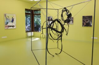 Ein Raum in einer Kunstgalerie mit gelben Wänden, in dem eine abstrakte Pferdeskulptur aus Draht und Riemen in einem Metallrahmen zu sehen ist sowie drei Mischtechnik-Gemälde an den Wänden hängen.